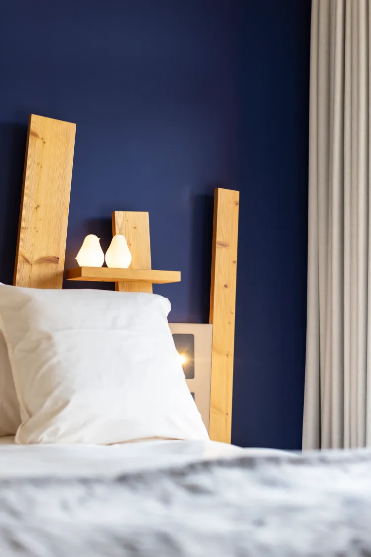 Un lit avec une tête de lit en bois clair et deux bougies allumées posées sur une étagère en bois, contre un mur bleu foncé