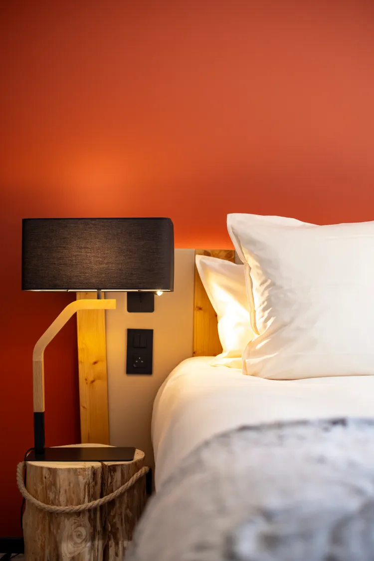 Lit avec une tête de lit en bois clair, deux oreillers blancs et une lampe de chevet à abat-jour noir sur une table de nuit en bois brut