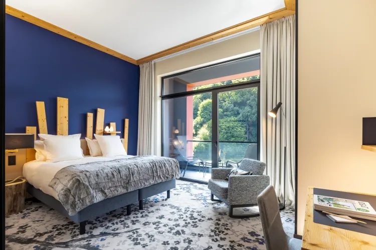 Chambre avec un lit double, une table de chevet, un fauteuil et une grande baie vitrée donnant sur une terrasse avec vue sur des arbres